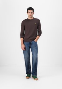 Pull en tricot marron à manches longues avec un emblème jaune, associé à un jean en denim bleu clair et des baskets vertes avec des semelles marron. Design simple.