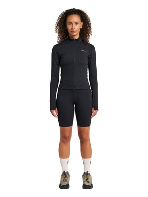 Kvinde med krøllet hår iført sort lynlåstrøje mærket "HALO", sorte cykelshorts, hvide sokker og beige vandresko, står oprejst.