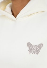 Cremfarbener Hoodie mit einem Schmetterlingsmotiv in Grau, das florale Details am Saum aufweist. Weicher Baumwollstoff mit lässigem Schnitt.