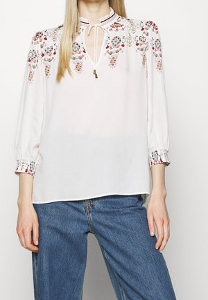 Mujer que lleva una blusa blanca con bordados florales rojos, escote con lazo y mangas de tres cuartos combinada con vaqueros azules.