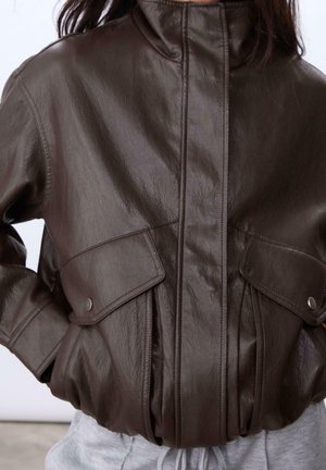 Persoon draagt een donkerbruine leren jas met klepzakken en drukkers, gecombineerd met lichtgrijze broek, handen in de zakken.