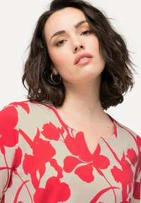 Blusa floreale beige con grandi motivi di fiori rossi. Presenta un design a scollo a V e maniche corte, realizzata in un tessuto leggero.