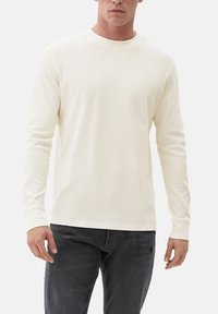 Camicia a maniche lunghe color crema con tessuto testurizzato, scollatura rotonda e polsini aderenti, abbinata a pantaloni grigi scuri.