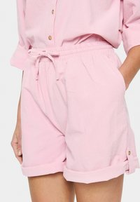 Pantaloni corti rosa chiaro con vita elastica, cordoncino, orli arrotolati, tasche laterali e una sottile trama. Dettagli con bottoni dorati sugli orli.