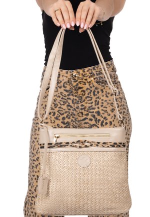 Frau trägt einen Rock mit Leopardenmuster und hält eine beige gewebte Handtasche mit Lederbesatz und Reißverschlusstasche an zwei Schulterriemen.