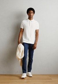 Polo shirt blanc à côtes, pantalon navy et sneakers blancs. Tissu léger drapé sur le bras gauche. Sol en bois, fond neutre.