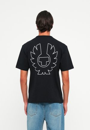 Man med svart t-shirt med vitt abstrakt vingemblem på ryggen och blå jeans, står framför en enfärgad vit bakgrund.