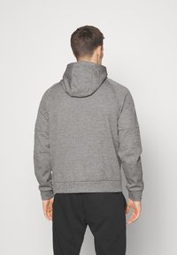 Sweat à capuche gris avec un tissu doux et texturé, doté d'une capuche, de manches raglan et d'un ourlet inférieur sans couture. Porté avec un pantalon de survêtement sombre.