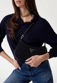 Borsa a spalla nera con una superficie liscia e strutturata. Caratterizzata da un logo triangolare dorato e una tracolla regolabile. Indossata sopra un maglione blu navy.