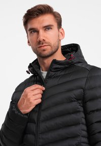 Schwarze Puffjacke mit Reißverschluss und Kapuze, mit gestepptem Design, elastischen Bündchen und kontrastierenden roten Akzenten an den Kordelzügen.