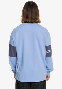 Osoba z włosami w kolorze brązowym do ramion, ubrana w jasnoniebieską koszulę z długim rękawem i ciemnoniebieskimi paskami na rękawach, odwrócona plecami.