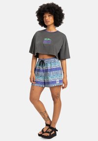 Grå kortermet t-skjorte med lilla kant og logo; blå mønstrede shorts med snøring; svarte platåsandaler; synlige tatoveringer.
