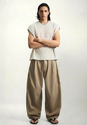 Junger Mann mit langen Haaren steht mit verschränkten Armen, trägt ein ärmelloses beiges Stricktop, weite beige Hosen, schwarze Flip-Flops und eine silberne Halskette.