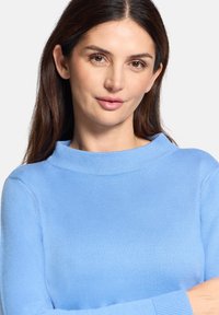 Kobieta o długich brązowych włosach, ubrana w jasnoniebieski sweter z dzianiny, patrząca przed siebie z neutralnym wyrazem twarzy, z założonymi skrzyżowanymi ramionami.