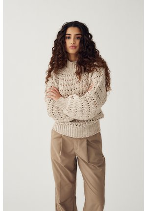 Jeune femme aux longs cheveux bouclés portant un pull en grosse maille beige et un pantalon ample marron, debout les bras croisés.