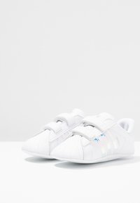Vita Adidas Superstar tofflor för småbarn med iriserande ränder och dubbel kardborreband på en vit yta och bakgrund.