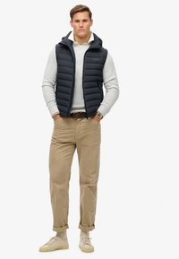 Marineblå puffer vest med hætte over en lys grå sweater, beige bukser og lyse sneakers, med et klassisk design og tekstureret stof.