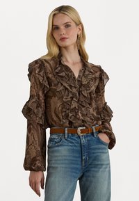 Brun paisley-mønstret bluse med volanger på ermene og kragen, kombinert med blå denimbukser og et brunt belte med en sølvspenne.