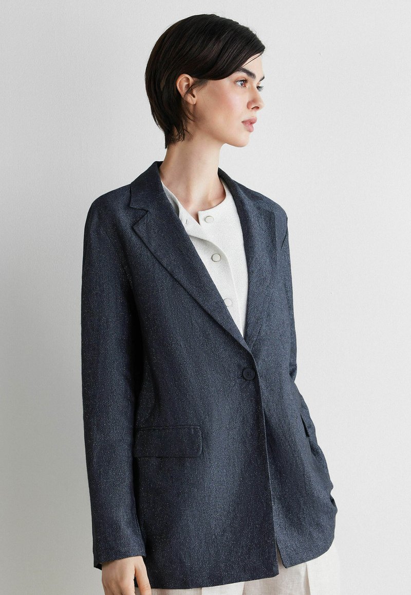 Femme aux cheveux courts et foncés portant un blazer texturé foncé sur un haut blanc boutonné, debout devant un fond clair uni.