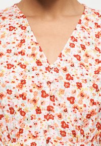 Hvit V-hals kjole med blomstertrykk i oransje, rødt og gult, med stofftrukne knapper ned foran og detaljer med innsvingt midje.