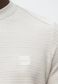 Gaiši bēšs džemperis ar apaļu kakla izgriezumu un teksturētu adījumu. Priekšpusē ir kvadrātveida logotipa etiķete nomierinošā tonī.