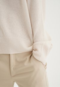 Pull en maille crème avec poignets et ourlet côtelés, associé à un pantalon beige taille haute, main posée de manière décontractée dans la poche du pantalon.