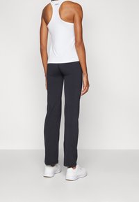 Bílé tílko s úzkými ramínky v kombinaci s tmavě šedými sportovními kalhotami, které mají hladkou texturu a přiléhavý střih. Bílé tenisky doplňují celý vzhled.