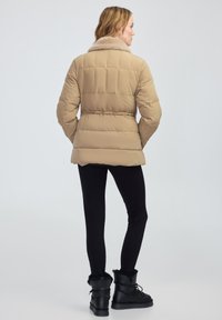 Beżowa kurtka puchowa z sztucznym futrem przy kołnierzu, pikowany wzór, dopasowana w talii, zestawiona z czarnymi legginsami i czarnymi botkami z futrzanym akcentem.