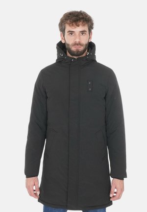 Parka nera con cappuccio, chiusura frontale con bottoni a pressione e tasche laterali. Presenta una superficie liscia, opaca e dettagli hardware minimi.