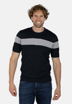 Hombre sonriente con pelo rizado, que lleva una camiseta negra de punto de manga corta con una franja horizontal gris y vaqueros azules, de pie frente a un fondo blanco.