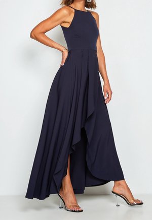 Žena předvádí tmavě modré šaty bez rukávů s delším zadním dílem a vzdušnou sukní, obutá do průhledných sandálů na vysokém podpatku, stojí před jednoduchým světlým pozadím.