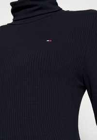 Mörkblå räfflad turtleneck-tröja med en åtsittande passform och liten broderad logotyp i röd, vit och blå på övre vänstra sidan.
