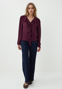 Blusa bordeaux con dettagli a ruches, maniche lunghe e scollatura a V. Abbinata a jeans a gamba larga blu scuro e ballerine intrecciate marroni.