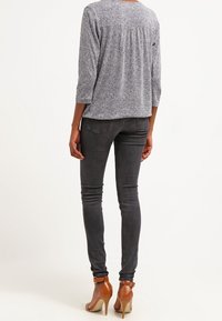 Frau, die eine graue gemusterte Bluse, dunkle graue enge Jeans und braune High-Heels mit Knöchelriemen trägt und von hinten steht.