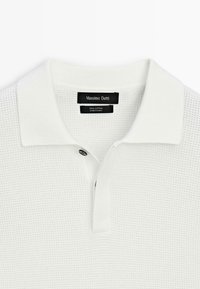 Polo in cotone bianco con motivo testurizzato, caratterizzata da un colletto classico e una chiusura con un solo bottone. Etichetta del marchio visibile sul colletto.