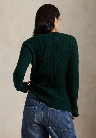 Donna con capelli scuri che indossa un maglione verde scuro a trecce e jeans blu, in piedi con una mano sui fianchi, davanti a uno sfondo beige.