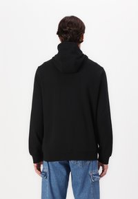 Sudadera con capucha negra de corte holgado, manga larga y puños acanalados. Textura suave con un diseño minimalista, combinada con jeans de color azul claro.