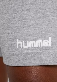 Close-up van grijze stoffen shorts met wit "hummel" merklogo boven de zoom op de dij van een persoon met een donkere huid.