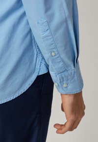 Light blue shirt fabric with long sleeves, featuring two buttons at the cuff and a rounded hem. Smooth texture with subtle stitching details.