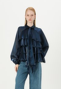 Blouse en chiffon superposée bleu marine avec des volants, un nœud au cou et de longues manches ballon, associée à un jean en denim bleu clair.
