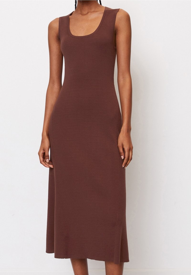 Femme portant une robe midi marron cintrée sans manches en maille côtelée, avec un col rond, debout devant un fond blanc uni.