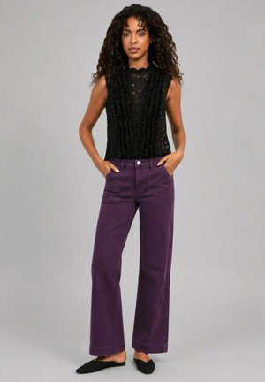 Femme debout, les mains dans les poches d’un pantalon large violet taille haute, portant un haut sans manches en dentelle noire et des chaussures plates noires pointues.