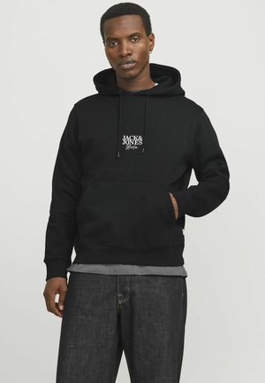 Hoodie noir en coton, avec une poche avant, un capuchon à cordon et un logo blanc sur la poitrine. Porte avec un pantalon en denim foncé.