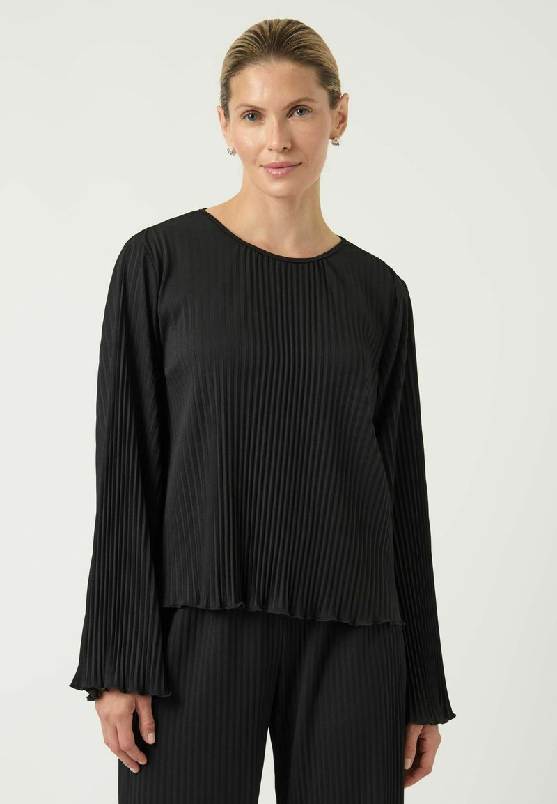 Svart plaggetopp med lange ermer og plissert stoff, rund hals, myk tekstur og utsvingte mansjetter, kombinert med matchende bukser.