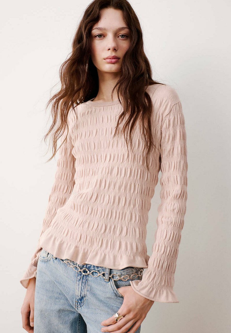 Jeune femme aux longs cheveux ondulés portant un haut rose clair texturé à manches longues, un jean bleu clair et une ceinture chaîne, debout contre un mur blanc.