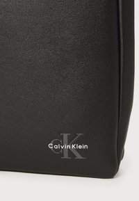 Borsa nera in pelle con superficie testurizzata, caratterizzata da un logo "CK" in argento e dalle parole "Calvin Klein" in bianco. Design rettangolare con linee pulite.
