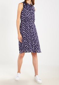 Modelka stojí ve bezrukávových fialkových šatech s vzorem a předním zapínáním na knoflíky, doplněná bílými teniskami, na světlém pozadí.