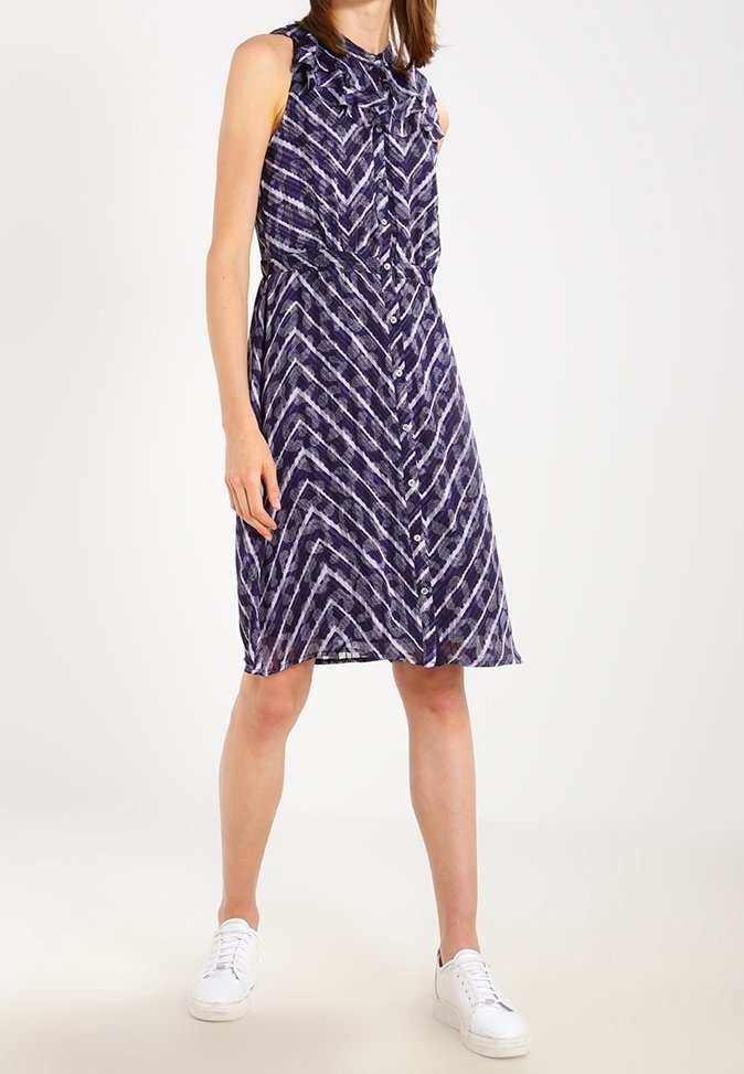 Modelka stojí ve bezrukávových fialkových šatech s vzorem a předním zapínáním na knoflíky, doplněná bílými teniskami, na světlém pozadí.