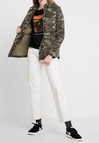 Chaqueta con estampado de camuflaje que presenta bolsillos en el pecho con botón, un zipper frontal y un corte holgado, combinada con unos vaqueros blancos cortos y zapatillas negras.