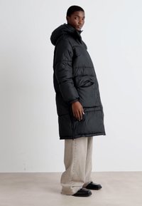 Black puffer coat with a hood, featuring a loose fit, vertical quilted pattern, and side pockets, worn over light-colored wide-leg pants.